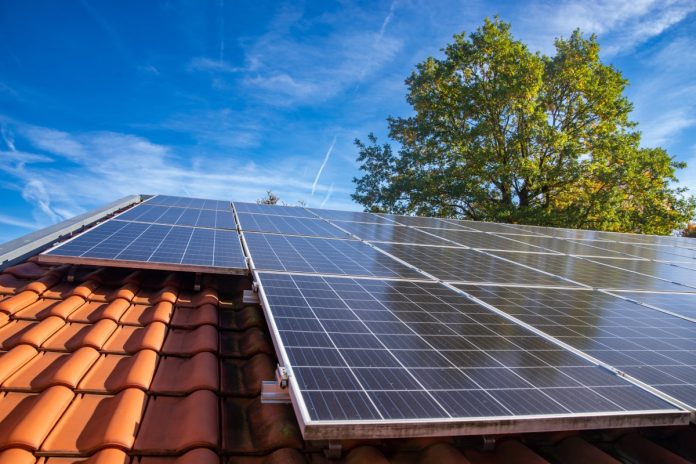 A close-up view shows solar panels on a home with orange slate shingles. A tree and the blue sky are in the background.