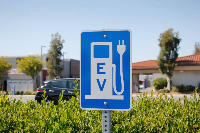 A blue and white sign signifying a charging station in a parking lot shows a cartoon "pump" with the initials "EV."