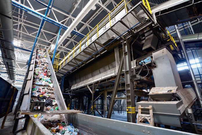 Crumpled plastic bottles and other packaging travel down a long conveyor belt at an industrial recycling facility.