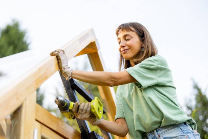 A young woman wearing a green tee shirt uses a yellow power drill to build a wooden structure while outside.