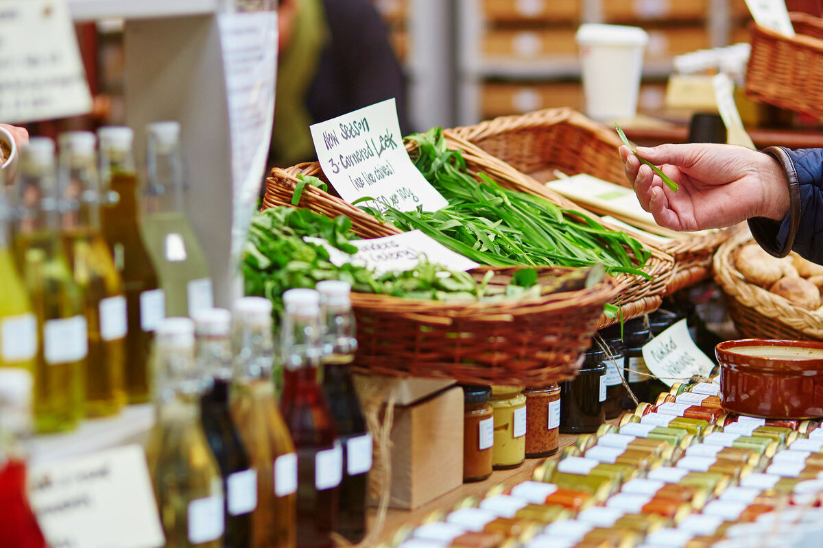 A person examining leeks for sale at a farmers market booth. There are also mushrooms and oils at the booth.