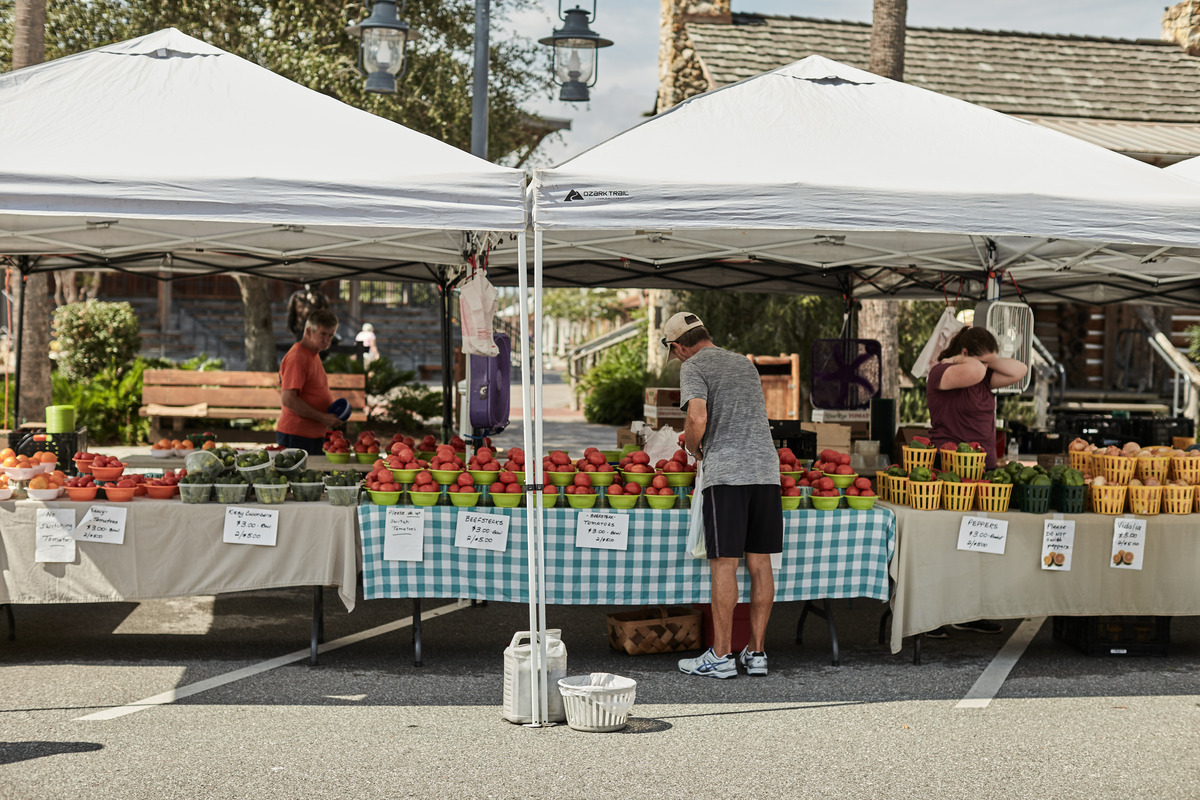 A male shopper stands at a farmers market booth looking at tomatoes for sale. Two booth attendants stand nearby.