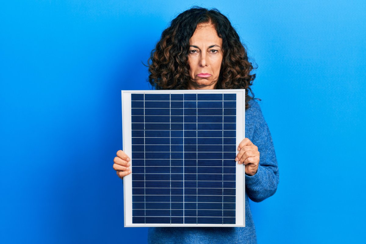A woman frowning while holding a small solar panel with both hands, standing against a blue background.