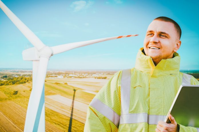 A man wearing a high-visibility jacket and holding a clipboard in front of a wind turbine beneath a blue sky.