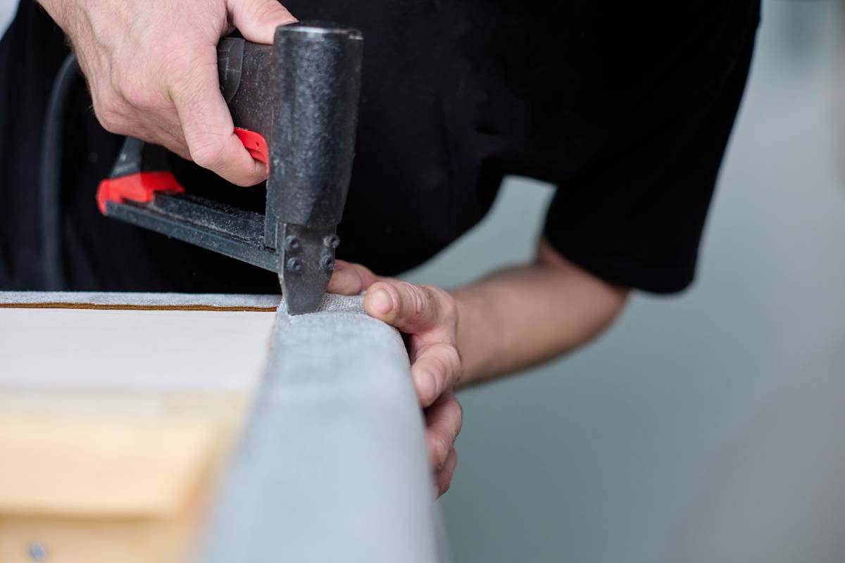 A person using a staple gun to attach fabric to a piece of furniture, securing upholstery along the edge.