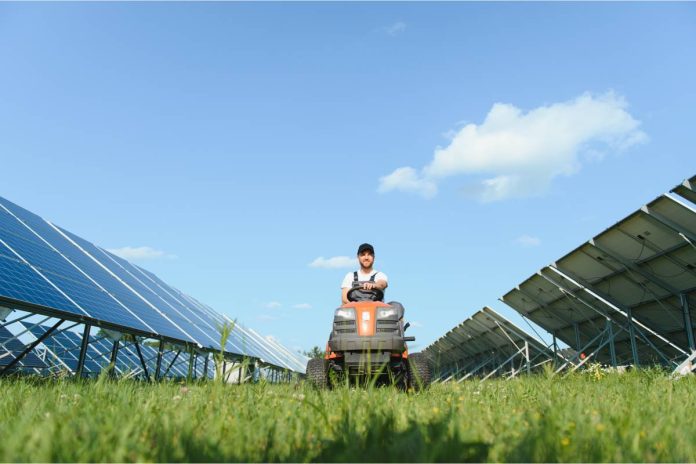 A young farmer driving a tractor down a stretch of grass between two rows of large blue solar panels on a farm.