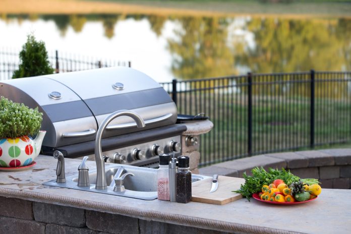 A side view of an outdoor kitchen with a sink and grill. A plate of vegetables and a cutting board rest on a countertop.
